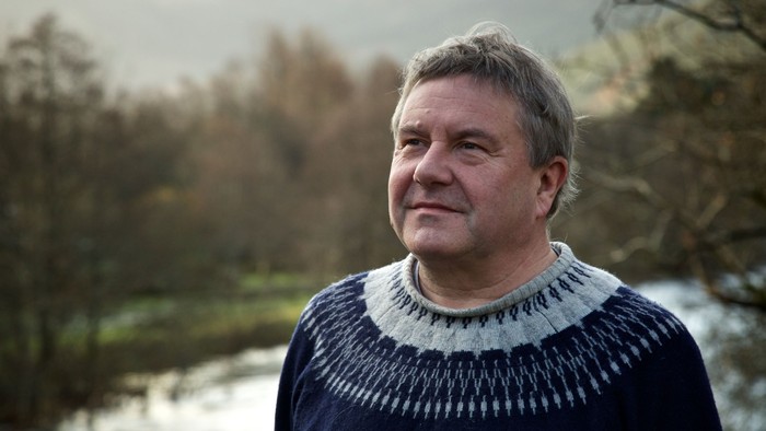 Middle-aged man in sweater standing outdoors near water.