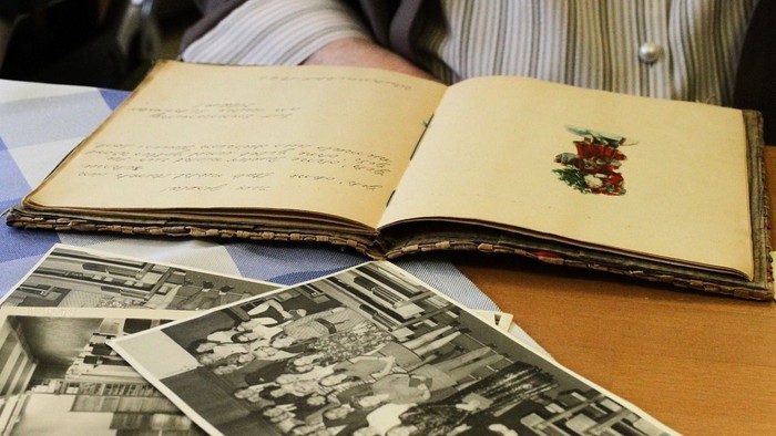 Ein aufgeschlagenes Buch mit handgeschriebenem Text und einer Stickerei, daneben liegen alte Schwarz-Weiß-Fotos