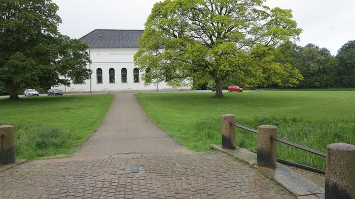 En stenvej leder op til et hvidt hus med arkade, omgivet af grønne træer og en græsplæne