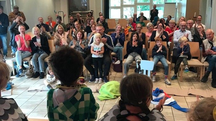 Eine Gruppe von Menschen, darunter auch Kinder, versammelt sich in einem Raum, während einige der Kinder vor ihnen auftreten oder etwas präsentieren.