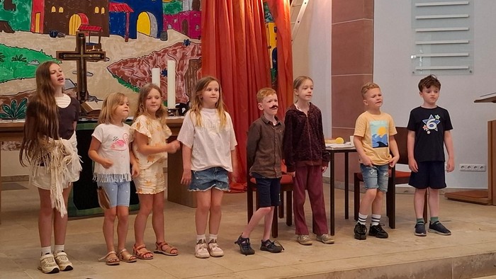 Eine Gruppe von Kindern steht auf der Bühne vor einer bunten Kulisse und führt wahrscheinlich ein Theaterstück auf oder singt.