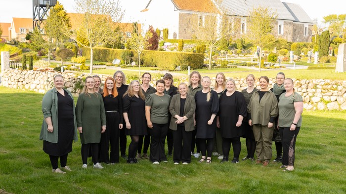En gruppe kvinder står sammen på en græsplæne foran en stor hytte med tårn og andre huse i baggrunden