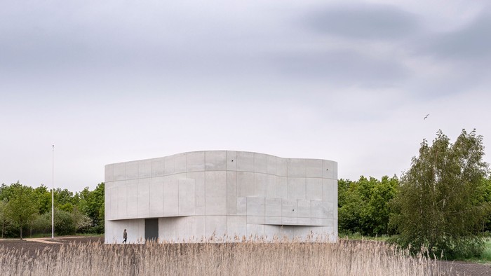 En moderne, kvadratisk betonbygning i en åben mark med høje græssarter og træer i baggrunden