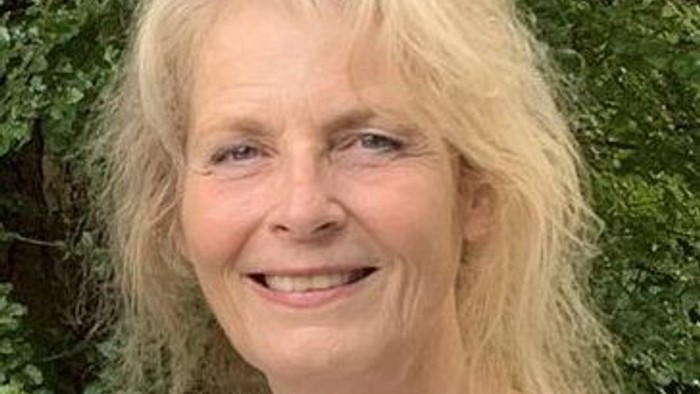 Kvinde med blondt hår, der smiler, stående udendørs foran en grøn, løvrig baggrund.