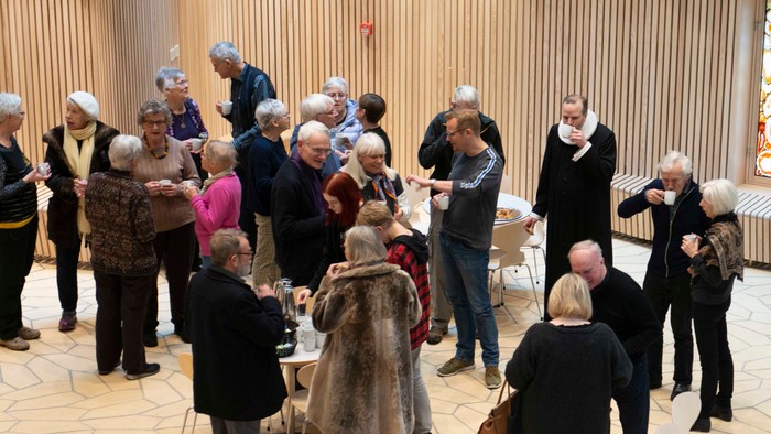 En gruppe ældre mennesker samles i et rum med høje kolonner og drikker kaffe.