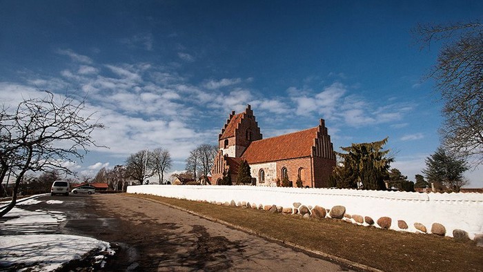 En rød kirke med spisse tårne og hvide murer, omgivet af træer og en klar blå himmel.
