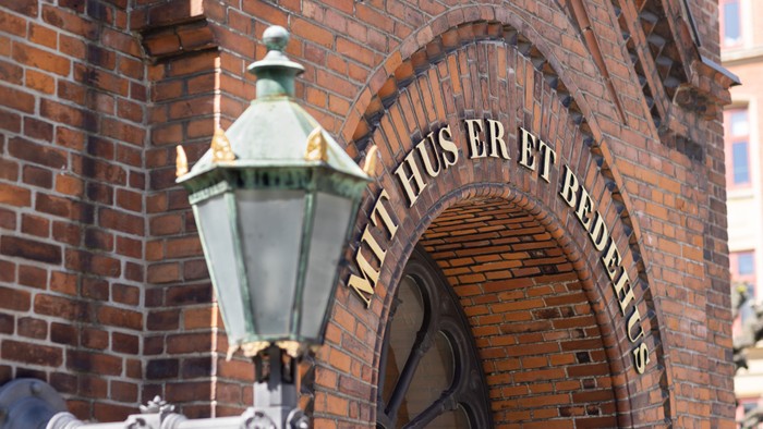 En lygte monteret på en mur af røde mursten med indskrift 'Mit hus er et bedste'.