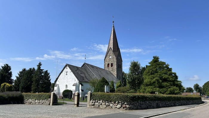 Hyggelig hvid kirke med et højt spir omgivet af en fredelig, anlagt landskabsområde under klare himmelstrøg.
