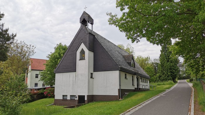 Kleine Kirche mit schwarzem Dach und weißer Fassade, umgeben von Bäumen und einem Weg.
