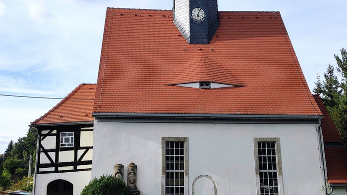 Kirchengebäude mit rotem Dach und hohem Turm mit Uhr und Wetterfahne