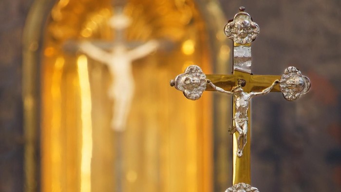 Verziertes goldenes Kruzifix mit aufwendigen Verzierungen, beleuchtet vor unscharfem Hintergrund.