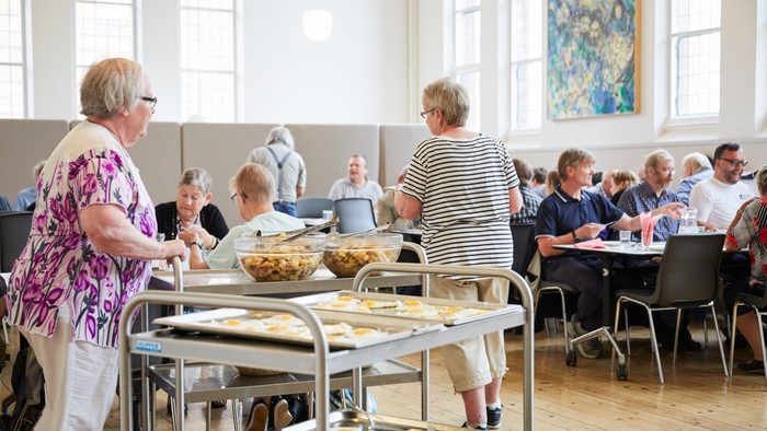 En gruppe ældre personer er samlet i en lys sal og serverer sig mad fra et buffet.