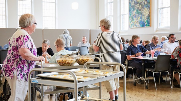 En gruppe ældre personer er samlet i en lys sal og serverer sig mad fra et buffet.
