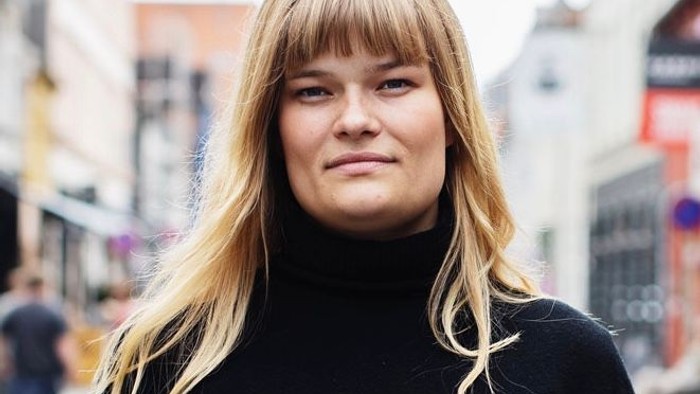 En ung kvinde med blondt hår og børste frisure står på en gade i en stor by.