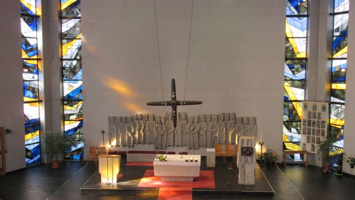 Moderner Kirchenraum der Allerheiligenkirche mit zentralem Steinaltar, einem schwebenden Bronzekreuz und einem großen Steinrelief von Heiligenfiguren im Hintergrund.