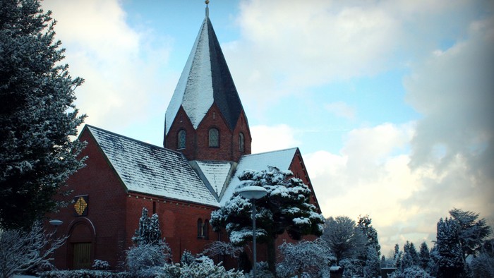En rød kirke med spiret dækket af sne under et bløligt himmel.