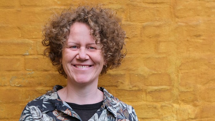 Kvinde med krøllet hår, der smiler foran en gul murstensvæg.