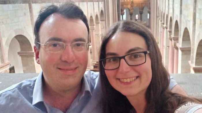 Zwei Personen, die in einem historischen architektonischen Ambiente mit Bögen und Säulen für ein Selfie lächeln.