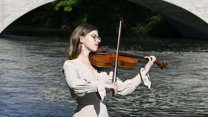 En kvinde i elegant påklædning spiller violin ved en rolig flod med en stenbro i baggrunden.