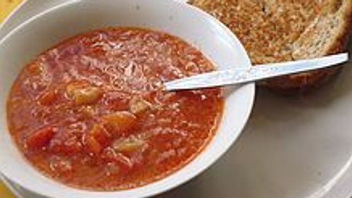 A bowl of tomato soup with a spoon next to a slice of toasted bread.