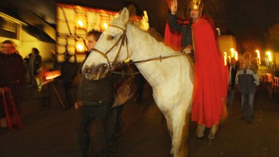 Ein Mann in rotem Umhang reitet auf einem weißen Pferd durch eine dunkle Straße, begleitet von mehreren Personen.