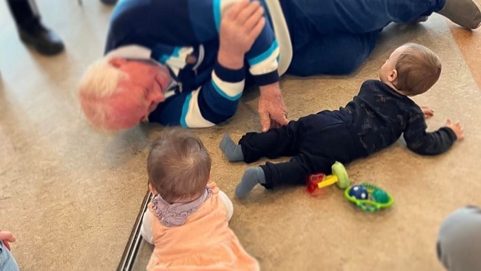 To børn spiller på gulv med legetøj, mens en voksen sitter ved siden af dem