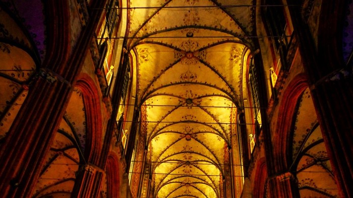Innenraum einer gotischen Kathedrale, in warmes, goldenes Licht getaucht, das die filigranen Gewölbedecken und Bögen hervorhebt.