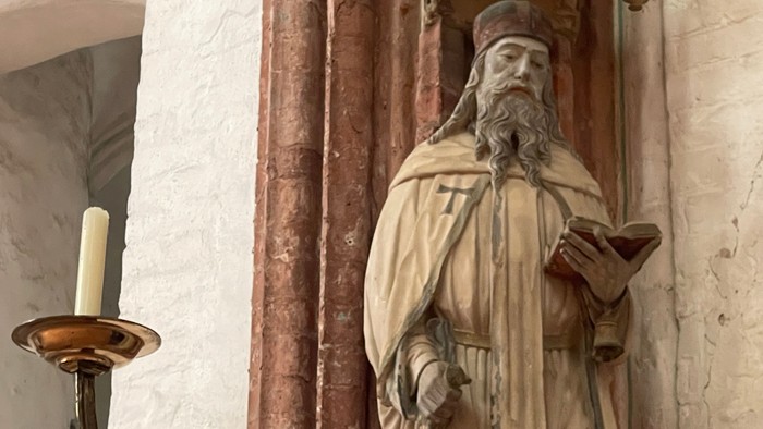 Statue einer bärtigen religiösen Figur, die ein Buch und einen Stab neben einem Kerzenhalter hält.