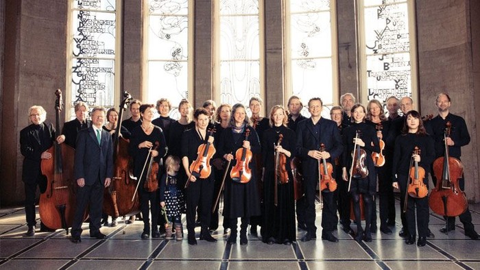 Ein professionelles Streichorchester steht gemeinsam in einem prächtigen Saal und hält verschiedene Instrumente.