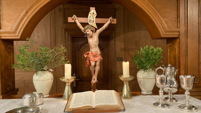 Das Bild zeigt eine religiöse Szene mit einem Kruzifix, Kerzen und einem offenen Buch auf einem Altar.