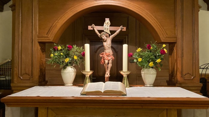 Das Bild zeigt einen Altar mit Kruzifix, Kerzen und Blumenarrangements.