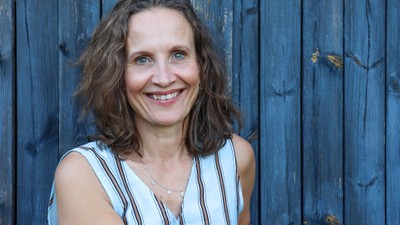 En glad kvinde i en stribet kjole læner sig op ad et træhegn og holder en gammel kuffert.