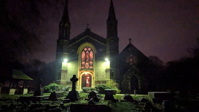 Kirchengebäude bei Nacht mit beleuchteten Fenstern und Grabsteinen im Vordergrund