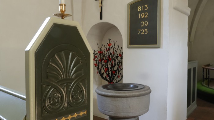 Elegant kirke døbefont med dekorativ plade, lysestage og blomsterarrangement ved siden af et kors.