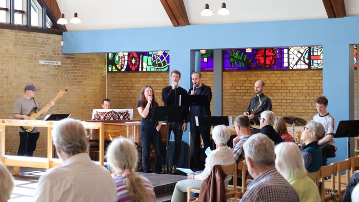 Fællesskabsarrangement med musikere, der optræder i en rummelig, lysfyldt sal med farvede glasvinduer.