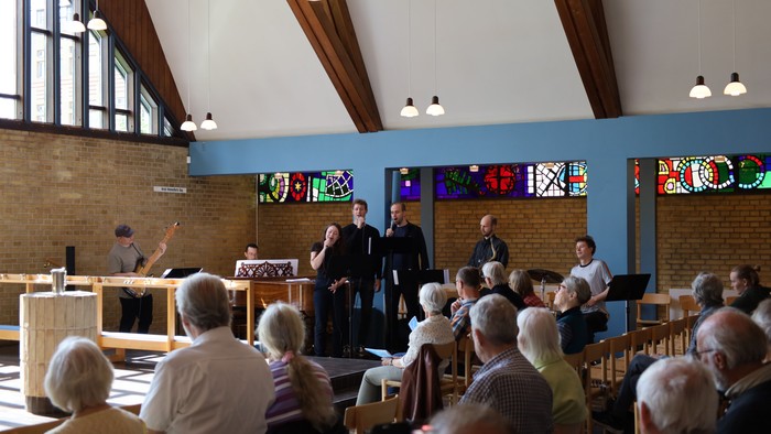 En gruppe samles i en kirke til en gudstjeneste, hvor præster taler, og menigheden sidder opmærksomt.