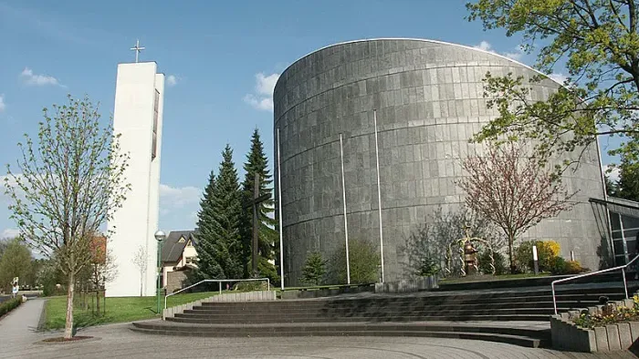 Moderne Kirche mit hohem Turm und gewölbter Fassade, umgeben von Bäumen und einem gepflasterten Vorplatz