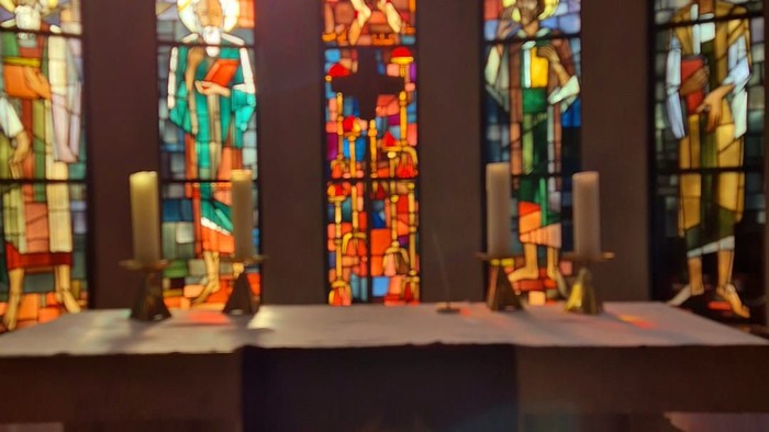 Das Bild zeigt das bunte Kirchenfenster. Es fällt warmes Sonnenlicht hindurch. Im Vordergrund ist der Altar mit 4 Kerzen zu sehen.