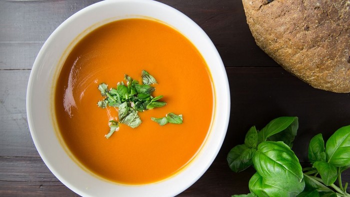 A bowl of orange soup garnished with green herbs, next to bread and basil leaves.