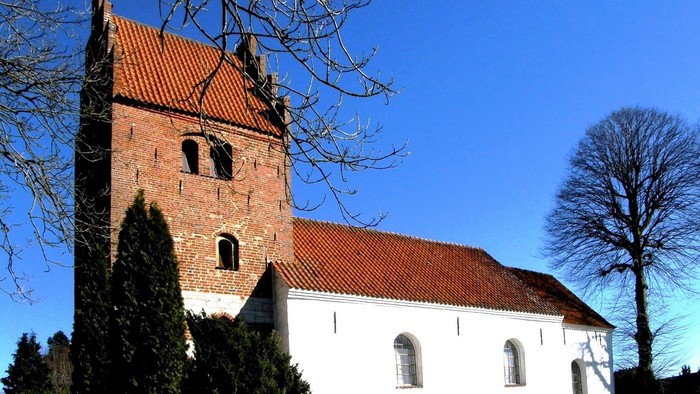 En hvid kirke med røde tag og tårn, omgivet af træer og buske under et klarblåt himmel.