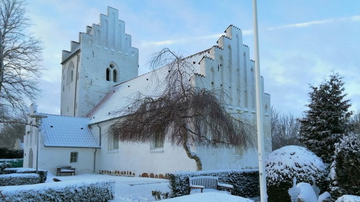 En snedækket kirke med tårn og en træ i forgrunden