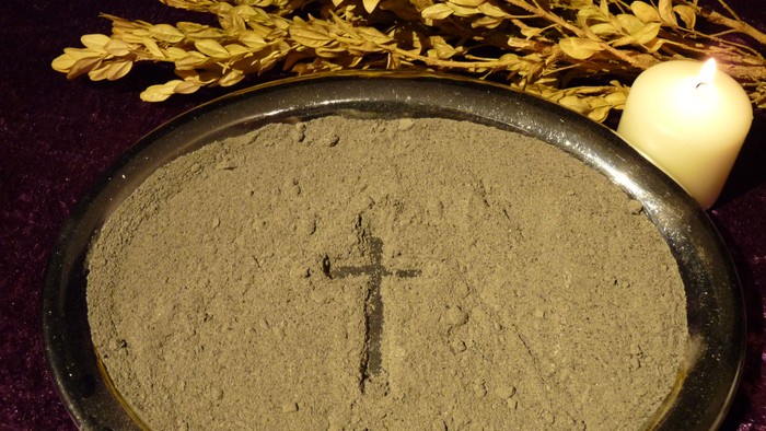 Auf einem Silbertablett liegt Asche. In der Mitte der Asche wurde ein Kreuz nachgezogen. Am oberen Bildrand sieht man noch einen alten Palmzweig aus Buchsbaum. Am oberen rechten Bildrand brennt eine kleine weiße Stumpenkerze.