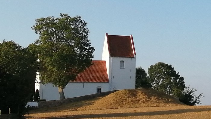 En lille hvid kirke med røde tag på en bakke med træer omkring