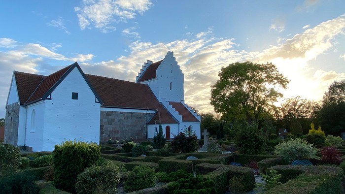 En hvid kirke med rødt tag og en have med grønne planter og træer ved solnedgang