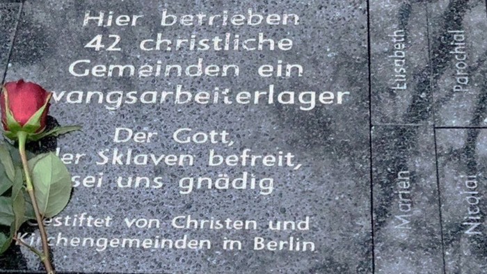 Ein roter Rosenblütenzweig liegt auf einem Grabstein mit Inschrift in deutscher Sprache.