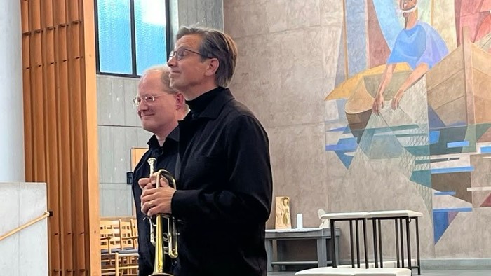 To mænd i en kirke holder messinginstrumenter og står nær farverige vægmalerier.