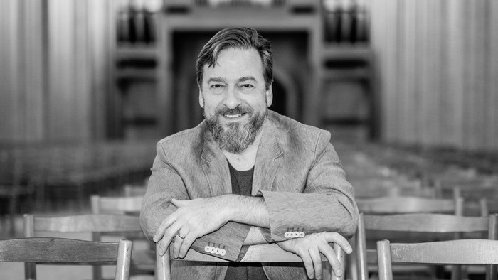 En skægget mand smiler varmt, mens han sidder på en stol i en indendørs omgivelse med arkitektoniske søjler.