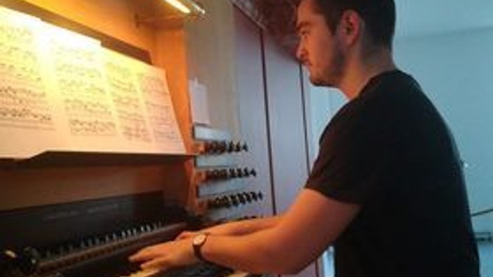 En mand spiller på et stort orgel med noder foran sig i et veloplyst rum.