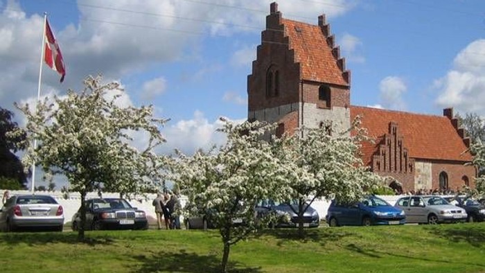 En kirke med røde tag og en blomstrende træ i forgrunden, med danske flag og biler i nærheden