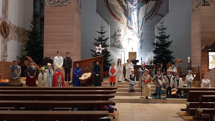 Eine Gruppe von Menschen versammelt sich in einer Kirche, einige stehen, andere sitzen auf Bänken, vorne befindet sich ein Weihnachtsbaum.
